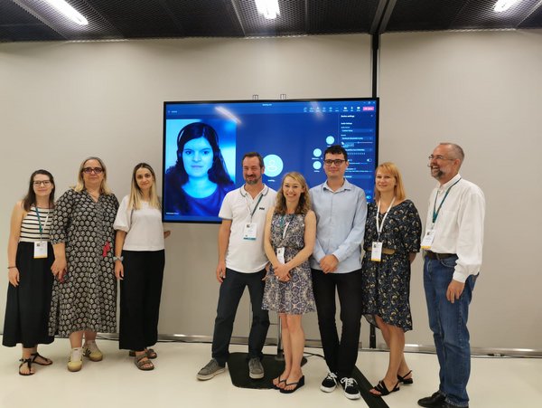 Eight people (5 women, 3 men) stand in front of a large screen with a profile picture of a woman on it